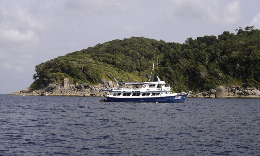 En blå og hvit safaribåt ankret foran en av Similan-øyene, med grå granittblokker i vannkanten under skogen