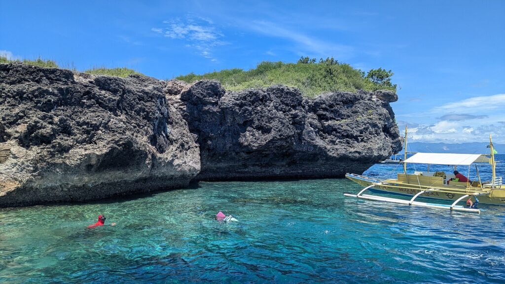 Den grå, uthulte kalksteinsklippen på Pescador Island, to svømmere med maske og snorkel og en gul banca i turkist vann