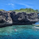Den grå, uthulte kalksteinsklippen på Pescador Island, to svømmere med maske og snorkel og en gul banca i turkist vann