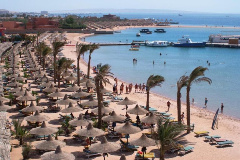 Stranden ved Giftun Azur Resort, med stråparasoller og palmer, og en brygge og båter i bakgrunnen