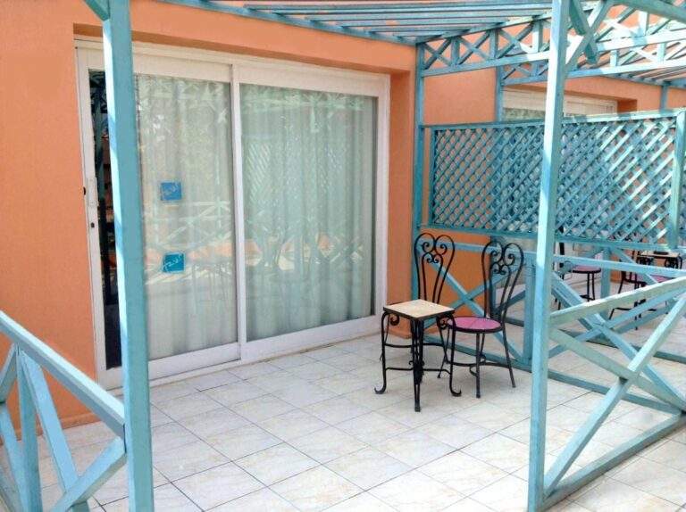En flislagt terrasse under en turkis pergola, med et lite bord og to stoler i smijern