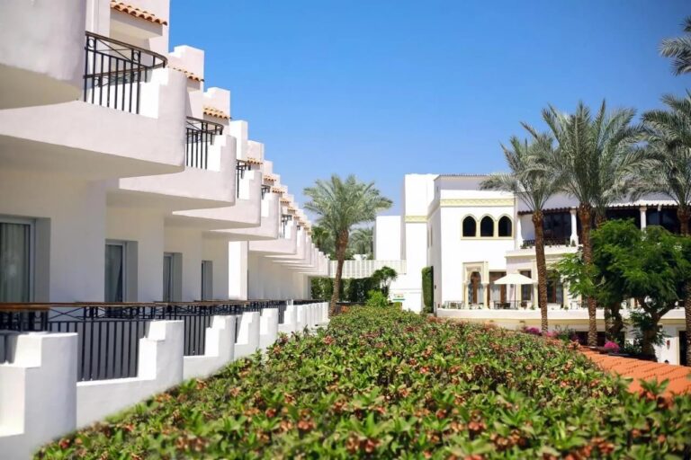 De hvite bygningene på JAZ Fanara Resort, med balkonger og palmer, bak en blomstrende hekk