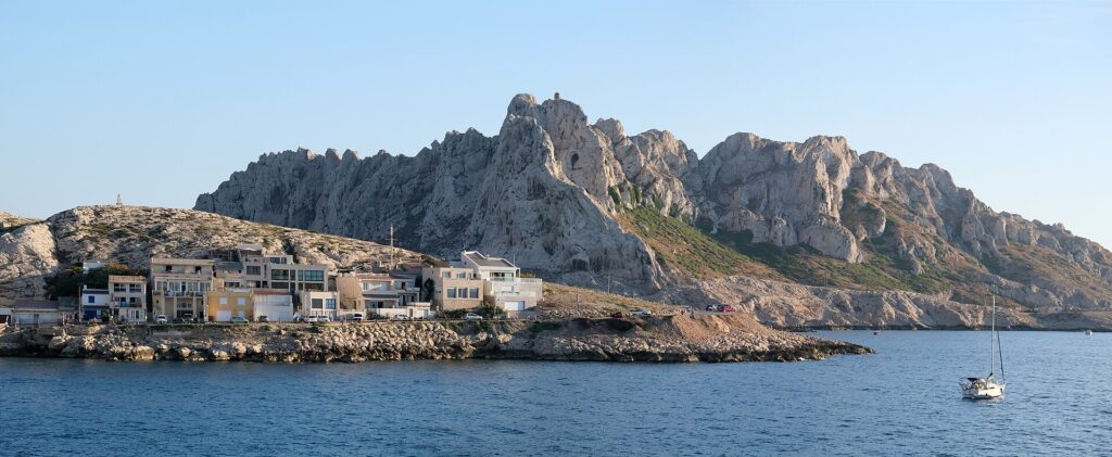 Husene i landsbyen Les Goudes på rekke helt nede ved en steinete kyst, foran en høy rygg av hvit kalkstein med taggete topper, under blå himmel, med en seilbåt for anker på stille sjø.