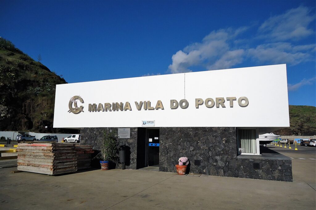 Mottaksbygningen i marinaen i Vila do Porto, et stort hvitt skilt med metallbokstaver på en vegg av svart basalt, under blå himmel, med en grønn klippe til venstre