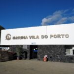 Mottaksbygningen i marinaen i Vila do Porto, et stort hvitt skilt med metallbokstaver på en vegg av svart basalt, under blå himmel, med en grønn klippe til venstre