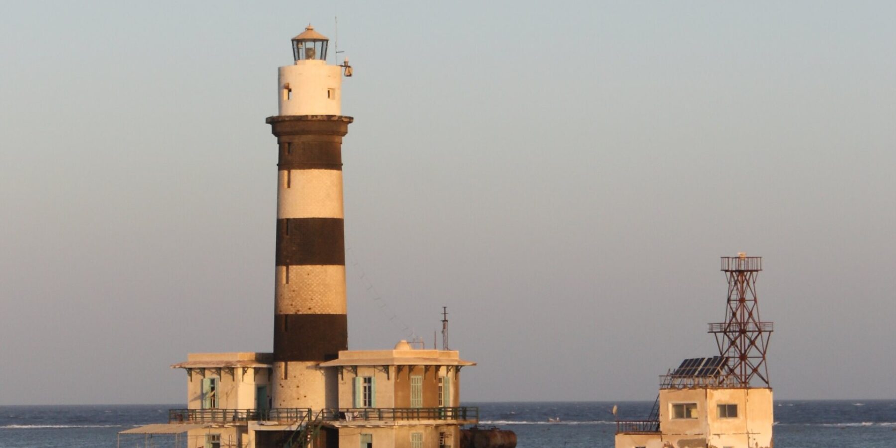 Fyrtårnet på Daedalus, et tårn med svarte og hvite striper på revet, med bygninger og en metallbrygge, i solnedgang