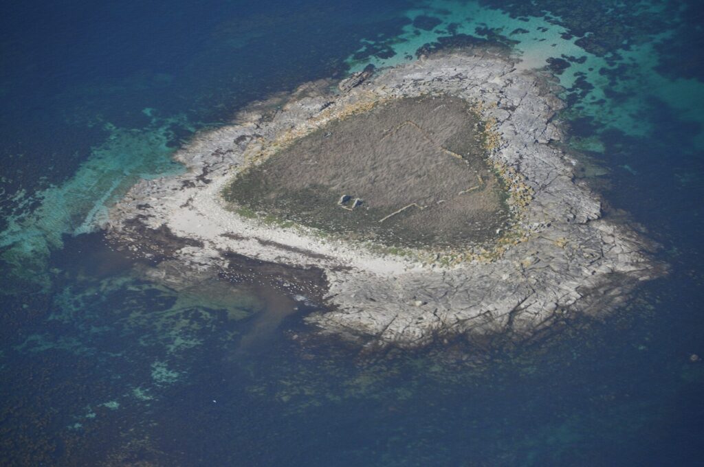 Flyfoto av en liten holme av grå granittheller, dekket av tørt gress i midten der du aner sammenraste steingjerder, kantet av en lys strand og omgitt av turkise grunner og dypblått vann.