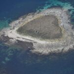 Flyfoto av en liten holme av grå granittheller, dekket av tørt gress i midten der du aner sammenraste steingjerder, kantet av en lys strand og omgitt av turkise grunner og dypblått vann.