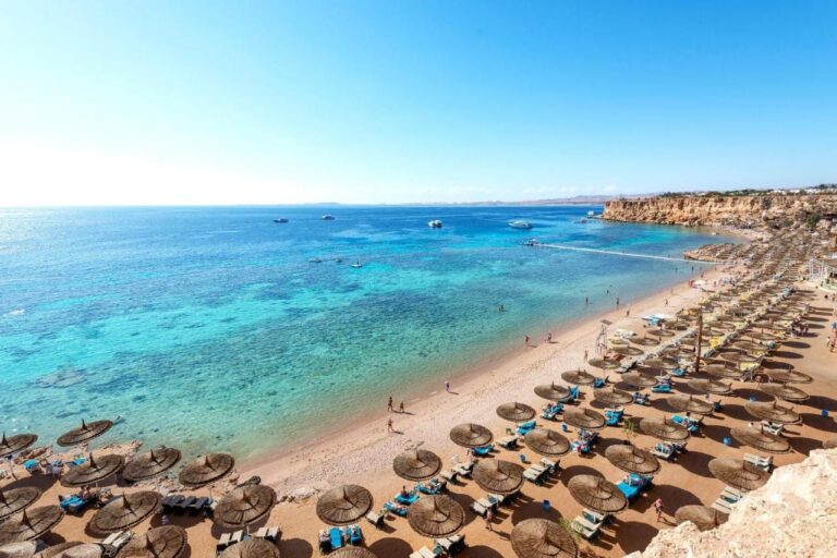 En lang sandstrand ved foten av en klippe, med parasoller av strå og en brygge ut i det turkise havet
