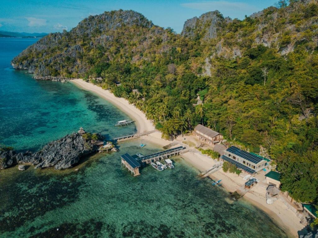 Sangat Island Dive Resort sett fra luften, med stranden, brygga og båtene, ved foten av klippene