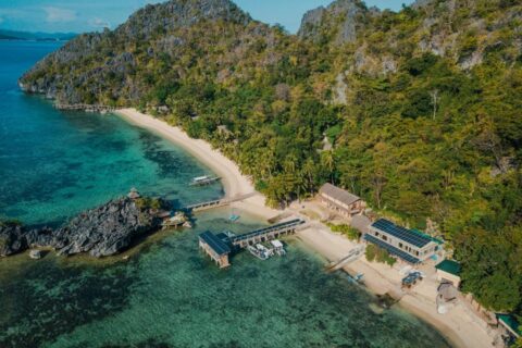 Sangat Island Dive Resort sett fra luften, med stranden, brygga og båtene, ved foten av klippene