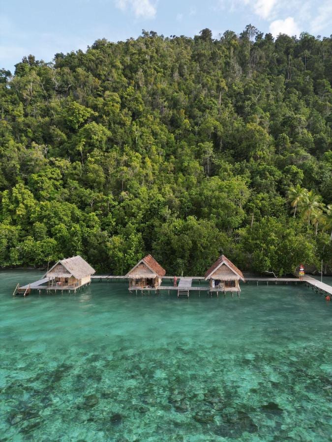 Tre bungalower på påler over turkist vann, ved foten av skogen
