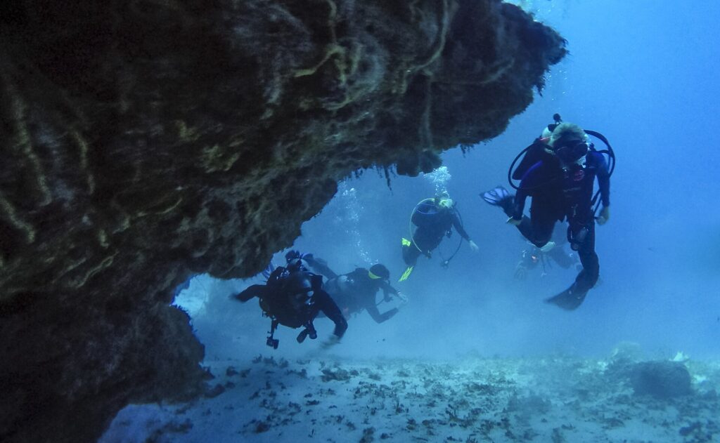Fire dykkere kommer ut av en koralltunnel over sandbunn på Cozumel, med steintaket over seg