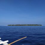 Den flate, skogkledde øya Balicasag i horisonten, med det hvite fyret og en stripe av lys sand, sett fra en bangka der de hvite utriggerne fyller forgrunnen, under en skyfri blå himmel.