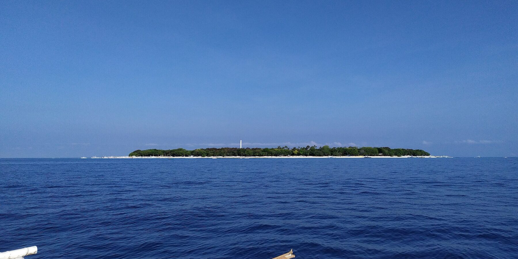 Den flate, skogkledde øya Balicasag i horisonten, med det hvite fyret og en stripe av lys sand, sett fra en bangka der de hvite utriggerne fyller forgrunnen, under en skyfri blå himmel.