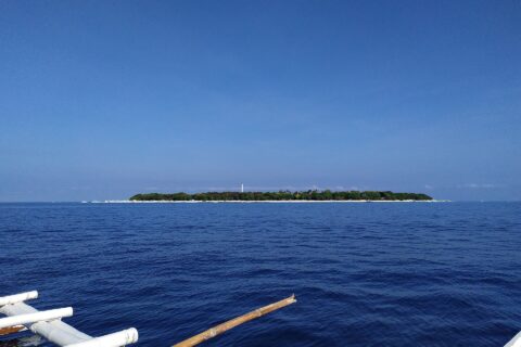 Den flate, skogkledde øya Balicasag i horisonten, med det hvite fyret og en stripe av lys sand, sett fra en bangka der de hvite utriggerne fyller forgrunnen, under en skyfri blå himmel.