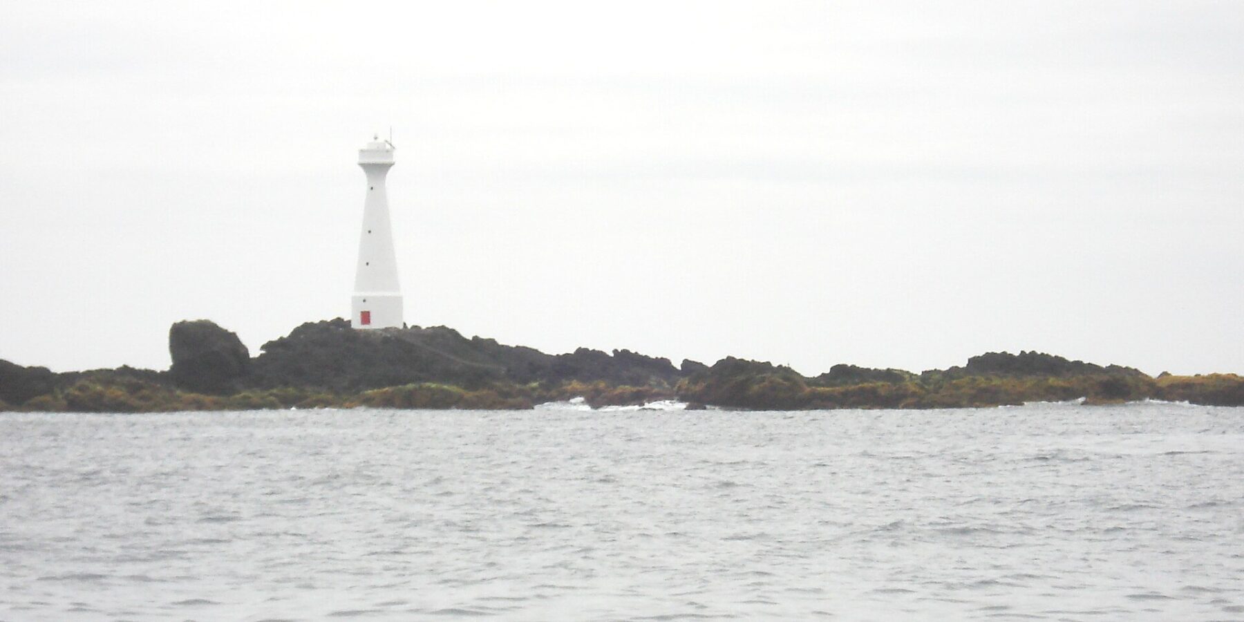 Et slankt hvitt fyr med rød dør står på den lave, steinete holmen Formigão, med mørke steiner kantet av brunalger, over et grått og urolig hav under overskyet himmel