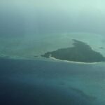 Øya Misali sett fra fly, en trekant av skog omkranset av en hvit strand, på en turkis revflate midt i et mørkeblått hav, i dis