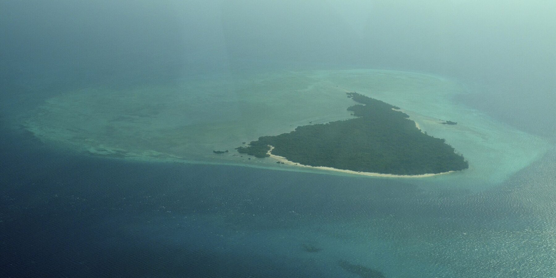 Øya Misali sett fra fly, en trekant av skog omkranset av en hvit strand, på en turkis revflate midt i et mørkeblått hav, i dis