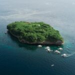 Flyfoto av Pescador Island, en klippe dekket av grønn vegetasjon med et hvitt fyr, omgitt av rundt ti dykkebancaer på mørkeblått hav