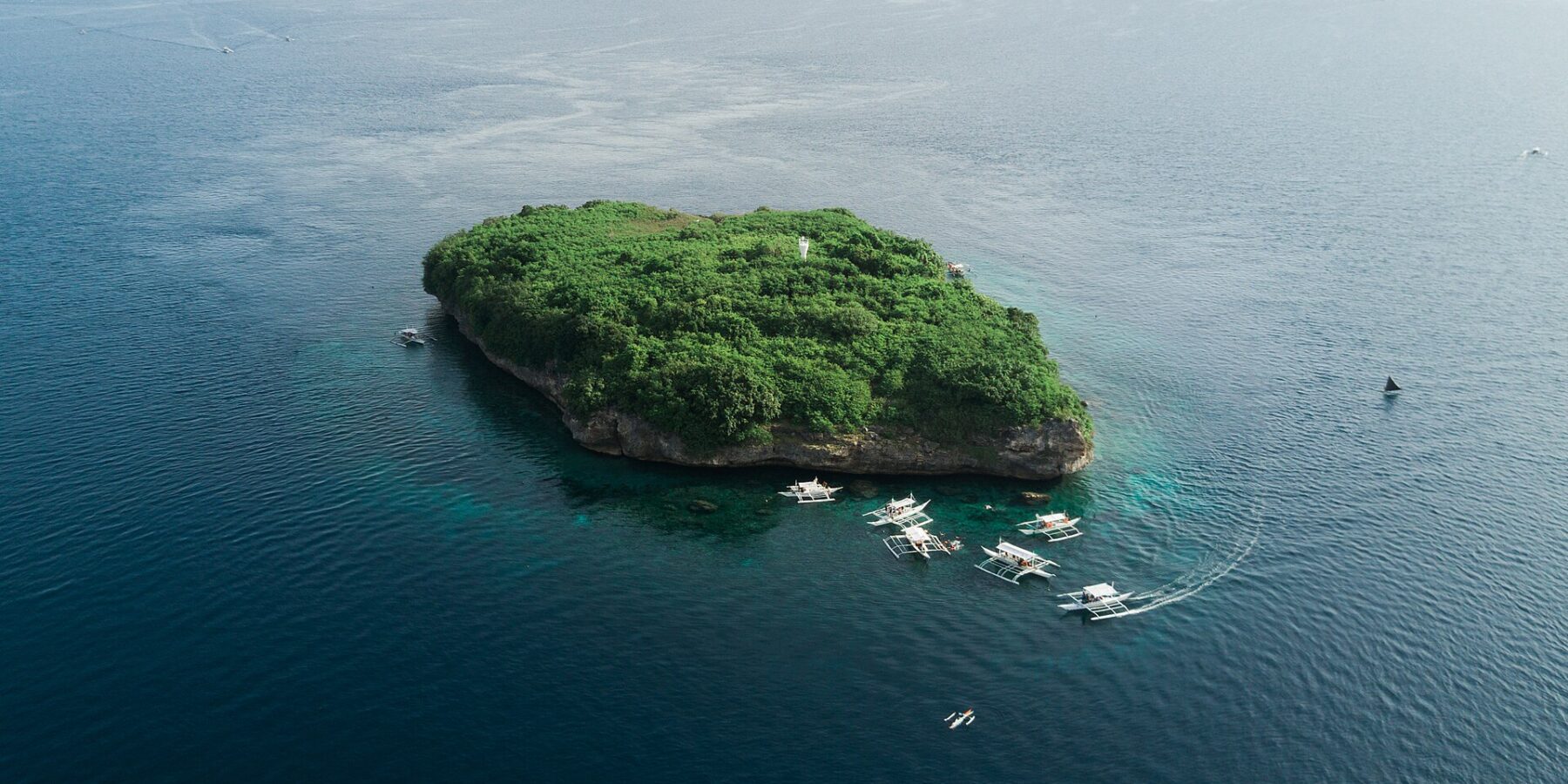 Flyfoto av Pescador Island, en klippe dekket av grønn vegetasjon med et hvitt fyr, omgitt av rundt ti dykkebancaer på mørkeblått hav
