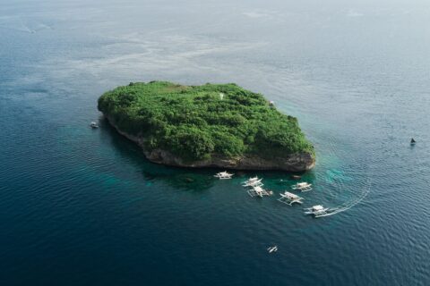 Flyfoto av Pescador Island, en klippe dekket av grønn vegetasjon med et hvitt fyr, omgitt av rundt ti dykkebancaer på mørkeblått hav