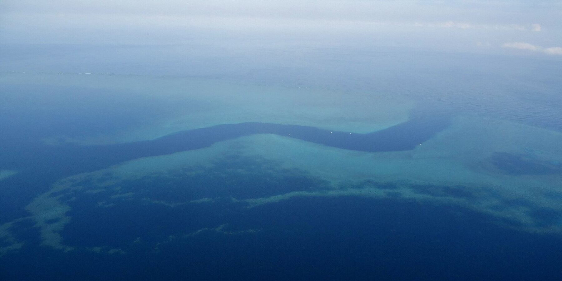 Flyfoto av en mørkeblå S-formet kanal som slynger seg mellom de turkise revflatene i barriererevet, med noen små hvite båter i passet