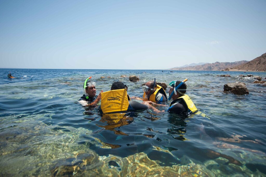 Svømmere i gule vester, med maske og snorkel, i det klare vannet i Blue Hole, med Sinai-fjellene bak