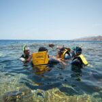 Svømmere i gule vester, med maske og snorkel, i det klare vannet i Blue Hole, med Sinai-fjellene bak